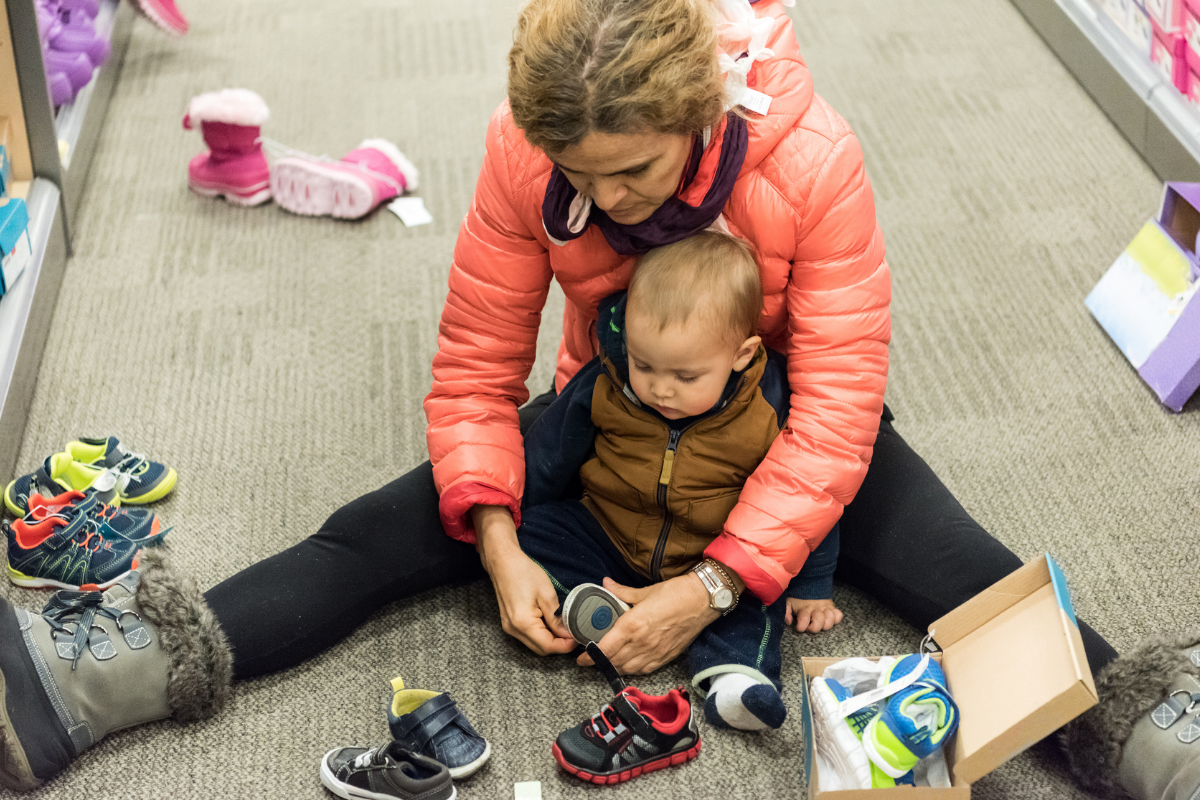 Keski-ikäinen valkoinen nainen lohenvärisessä toppatakissa istuu kenkäkaupan lattialla ja hän laittaa häntä vasten nojaavalle istuvalle vauvalle tarralenkkareita.